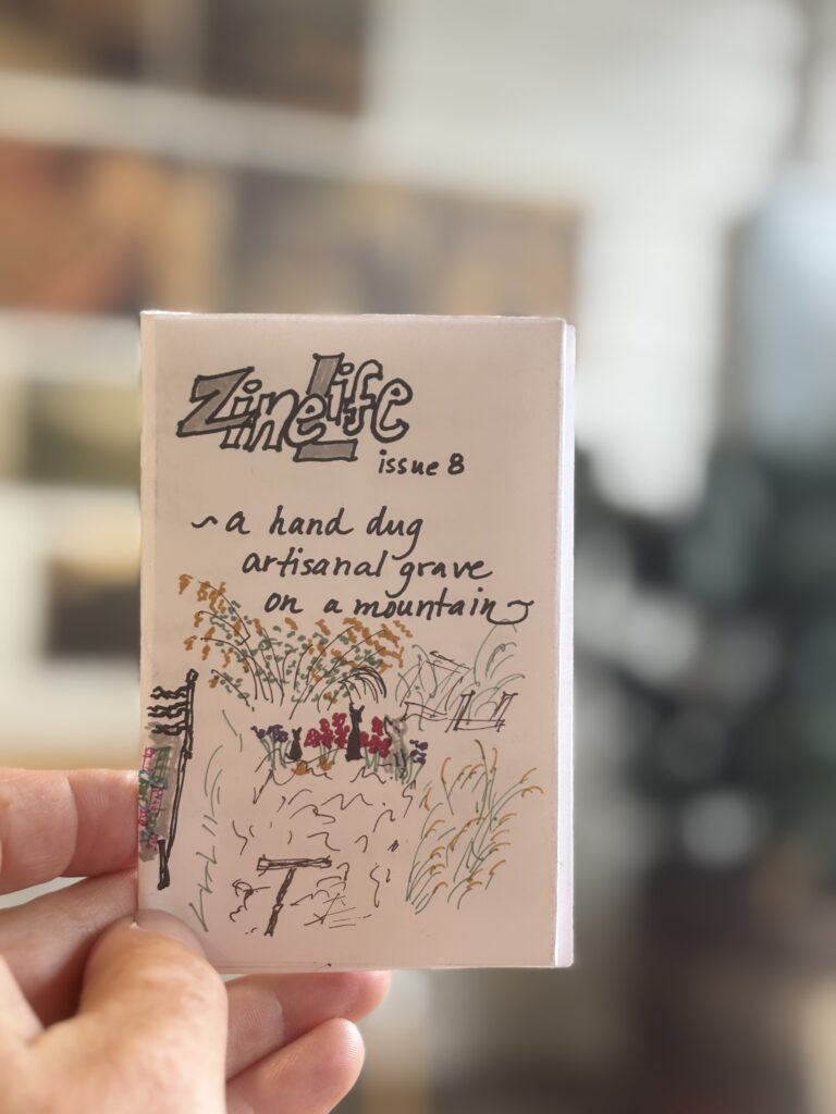 a hand drawn zine in a hand. zine life issue 8. it says a hand dug artisanal grave on a mountain and has a drawing of a grave next to a bed with a T on it